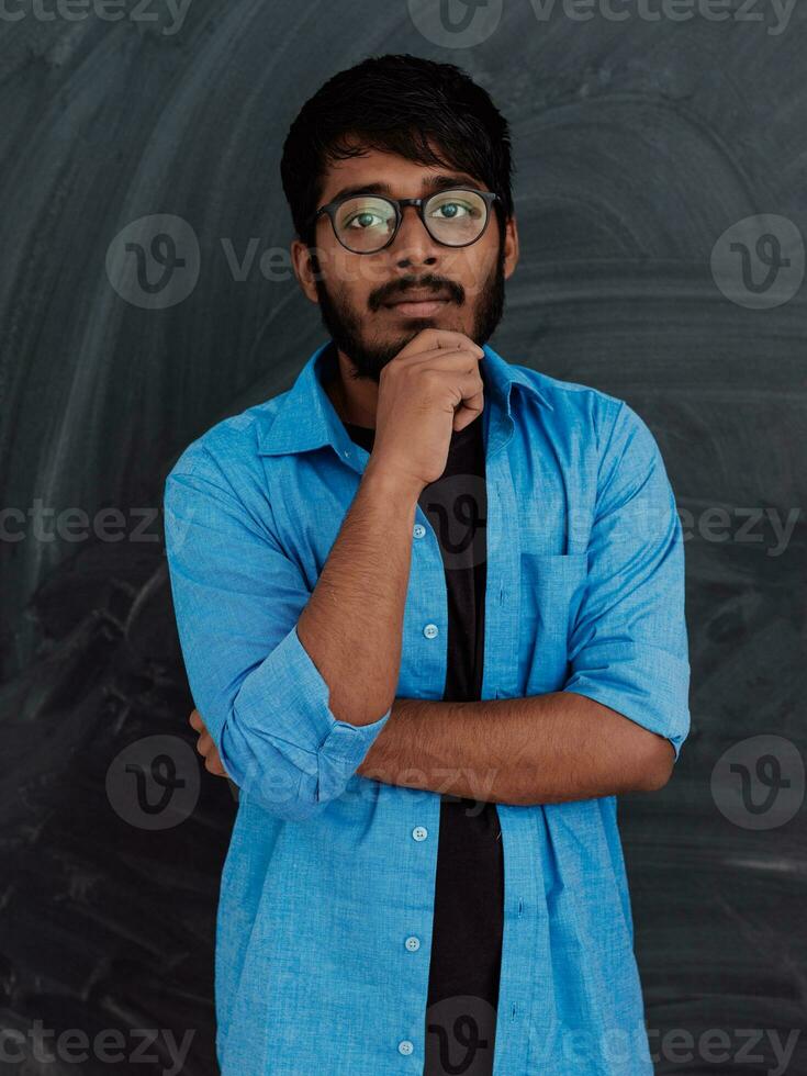 uma jovem indiano homem dentro uma azul camisa e óculos poses pensativamente dentro frente do escola borda foto