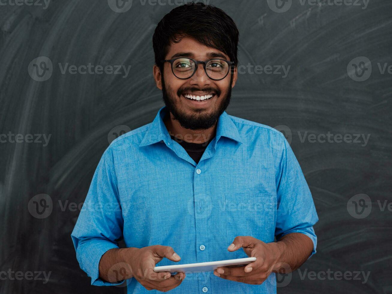 indiano sorridente jovem aluna dentro azul camisa e óculos usando tábua, Smartphone e posando em escola quadro-negro fundo foto