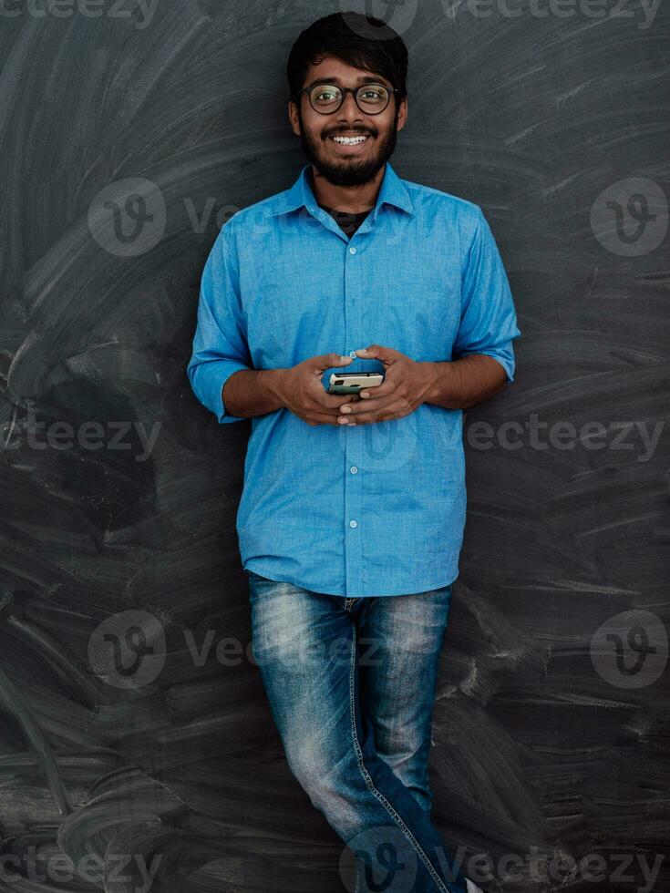 indiano sorridente jovem aluna dentro azul camisa e óculos usando Smartphone e posando em escola quadro-negro fundo foto
