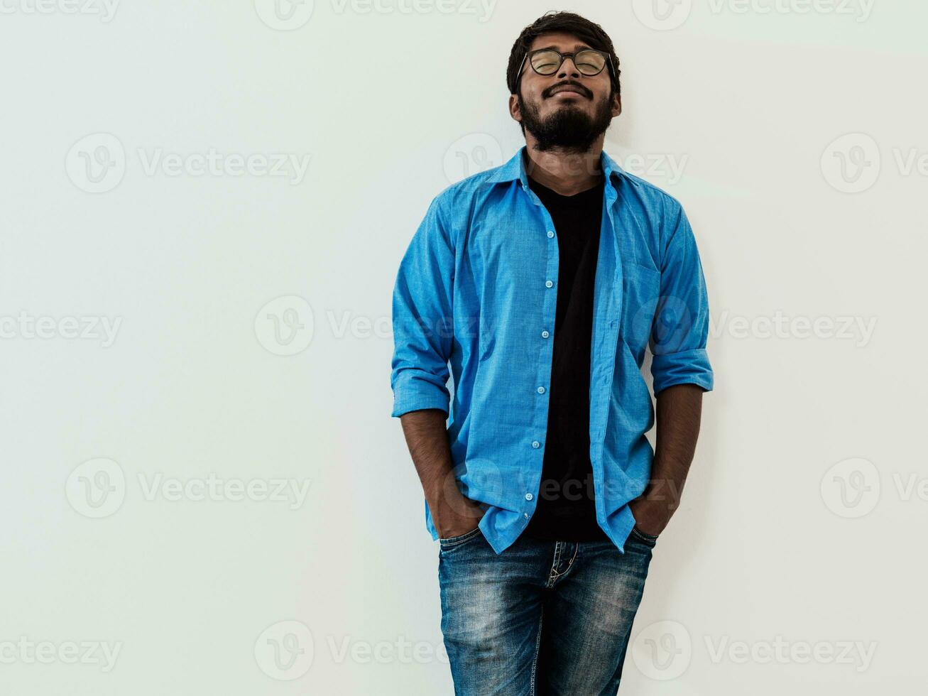 indiano sorridente jovem homem com azul camisa e óculos posando em cinzento fundo foto