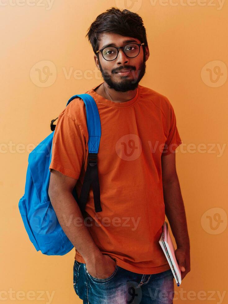 indiano aluna com azul mochila, óculos e caderno posando em laranja fundo. a conceito do Educação e escolaridade. Tempo para ir costas para escola foto