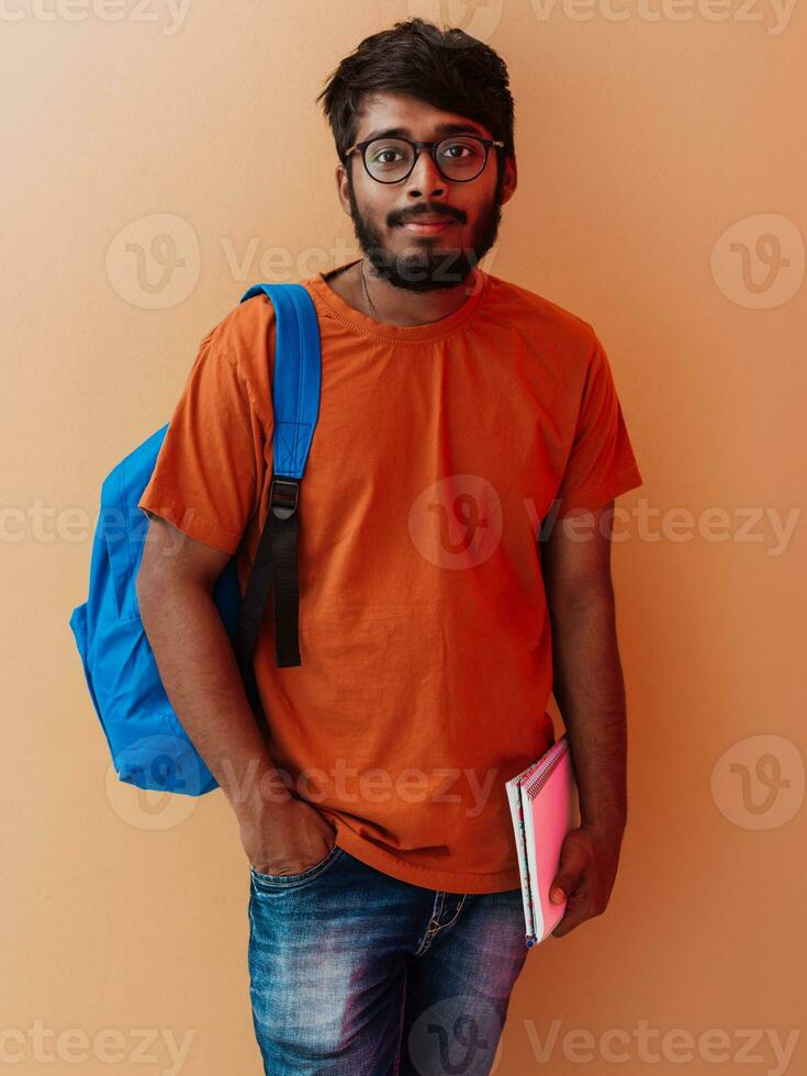 indiano aluna com azul mochila, óculos e caderno posando em laranja fundo. a conceito do Educação e escolaridade. Tempo para ir costas para escola foto