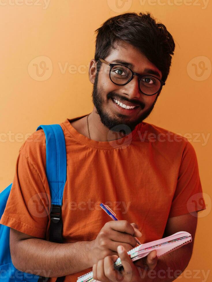 indiano aluna com azul mochila, óculos e caderno posando em laranja fundo. a conceito do Educação e escolaridade. Tempo para ir costas para escola foto