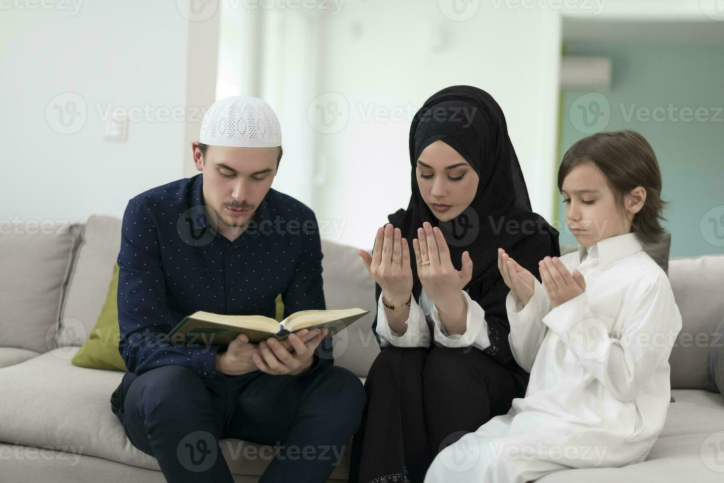 tradicional muçulmano família pais com crianças lendo Alcorão e Rezar juntos em a sofá antes iftar jantar durante uma Ramadã festa às casa foto