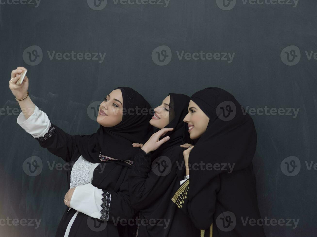 grupo do jovem muçulmano mulheres dentro elegante vestir com hijab usando Smartphone enquanto levando selfie cenário dentro frente do Preto fundo foto