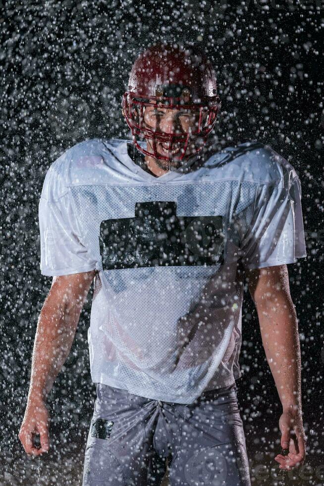 americano futebol campo solitário atleta Guerreiro em pé em uma campo detém dele capacete e pronto para jogar. jogador preparando para correr, ataque e Ponto aterragem. chuvoso noite com dramático névoa, azul luz foto