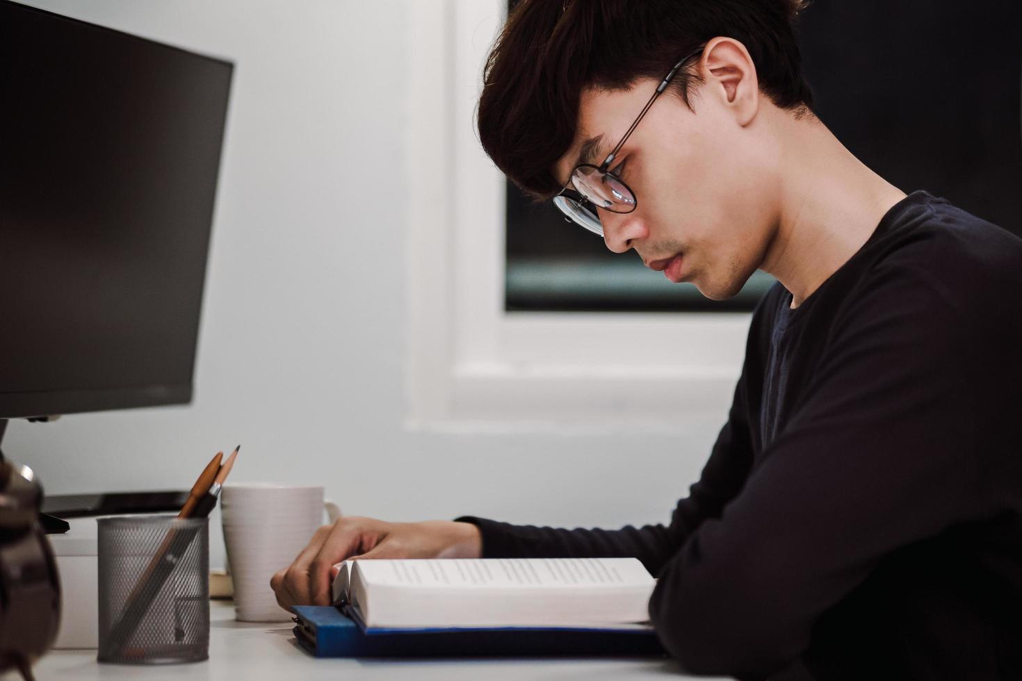 jovem bonito asiático lendo livro na mesa de trabalho tarde da noite foto