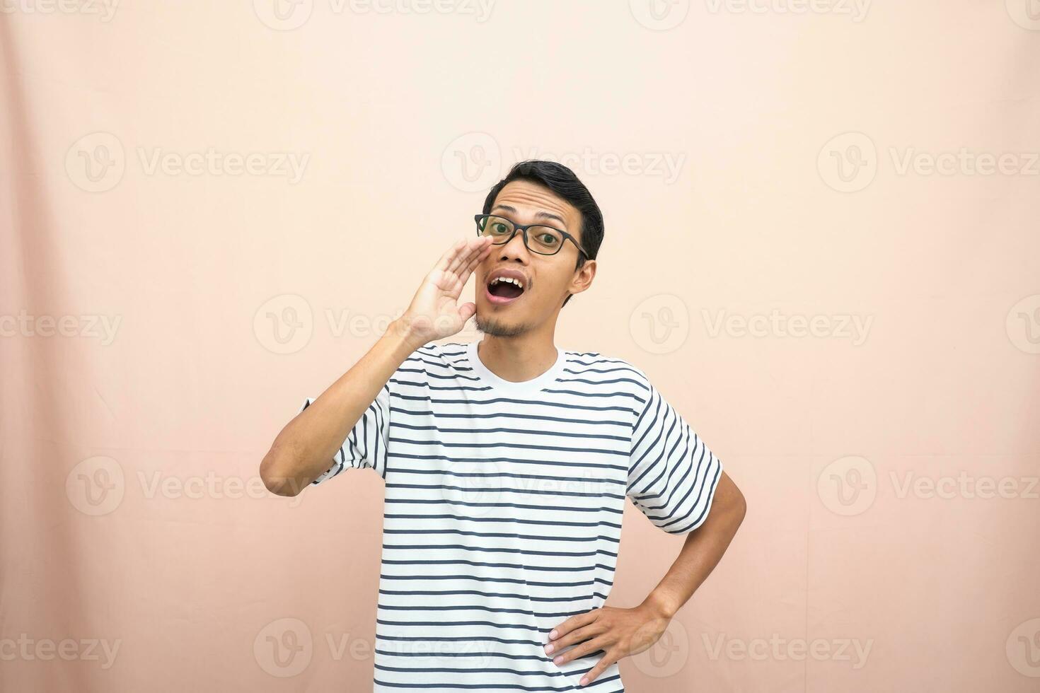 ásia homem dentro óculos vestindo casual listrado camisa, gritando pose com mão perto boca, dando Informação. isolado bege fundo. foto