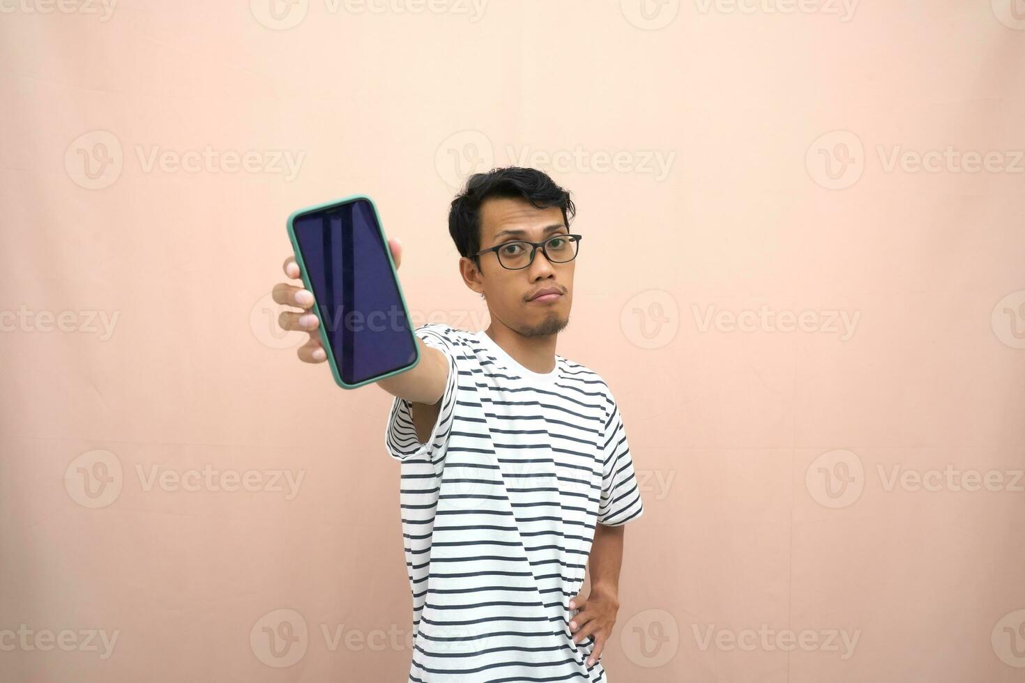 retrato do a ásia homem vestindo óculos vestindo uma casual listrado camiseta. mostra Smartphone tela com feliz, surpreso, feliz expressão, para promoção. isolado bege fundo. foto