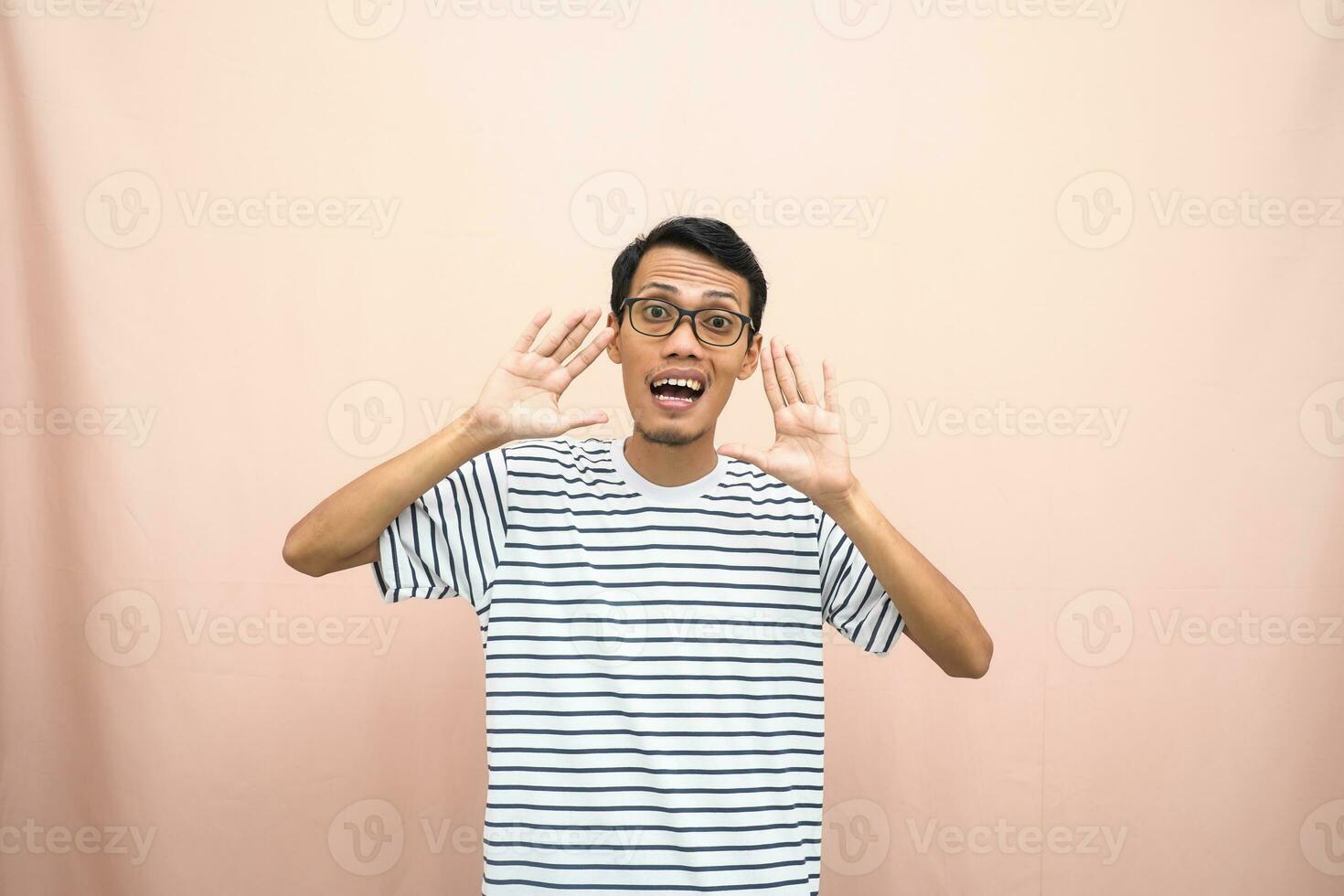 ásia homem dentro óculos vestindo casual listrado camisa, gritando pose com mão perto boca, dando Informação. isolado bege fundo. foto