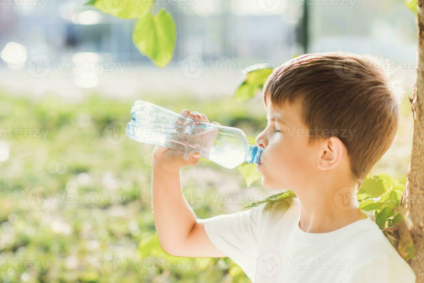 uma lindo criança sentado em a Relva bebidas água a partir de uma garrafa dentro a verão às pôr do sol. Garoto sacia dele sede em uma quente dia foto