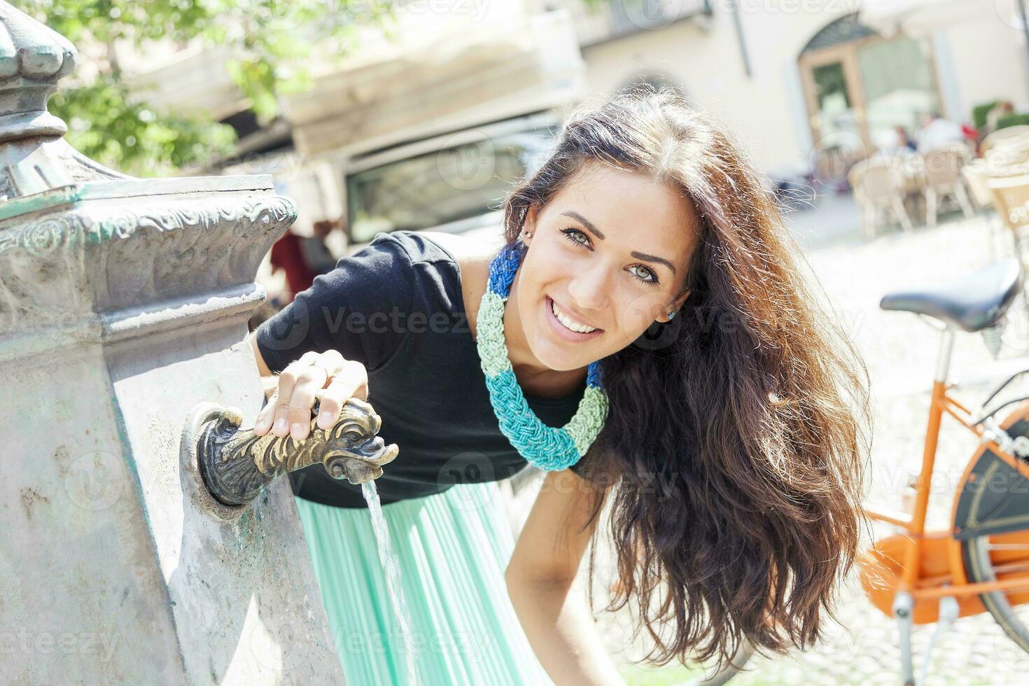 retrato do bonita menina bebidas água a partir de fonte dentro verão cidade foto