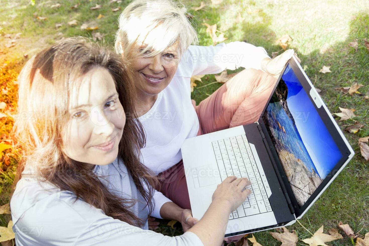 bonita mãe e filha consultar a computador portátil debaixo uma árvore dentro outono foto