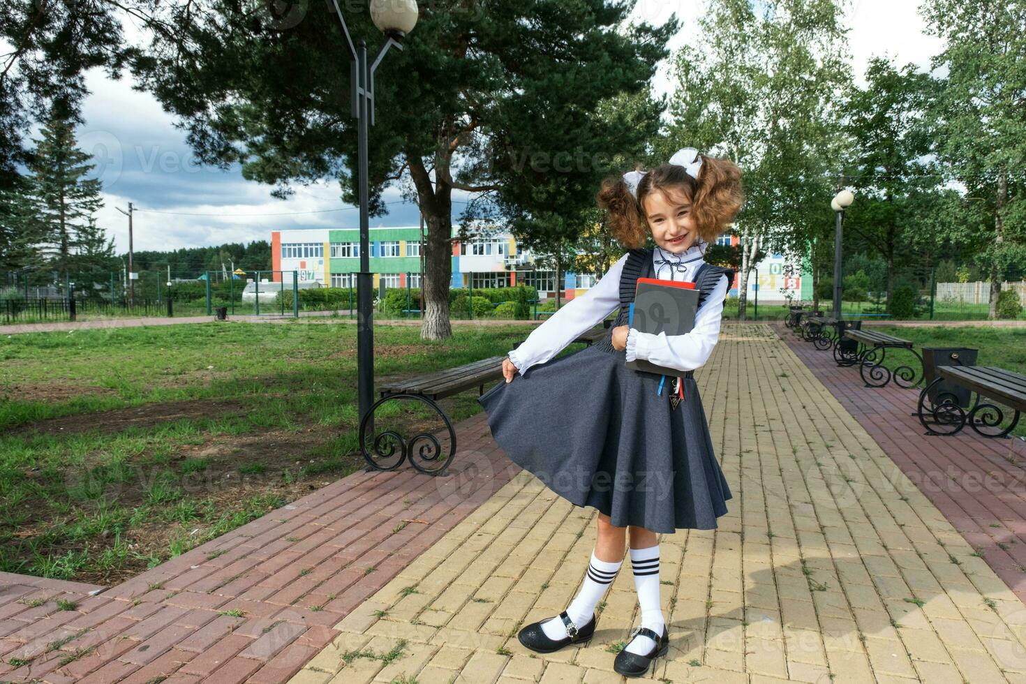 menina com mochila, escola uniforme com branco arcos e pilha do livros perto escola. costas para escola, feliz aluno, pesado livros didáticos. Educação, primário escola Aulas, setembro 1 foto