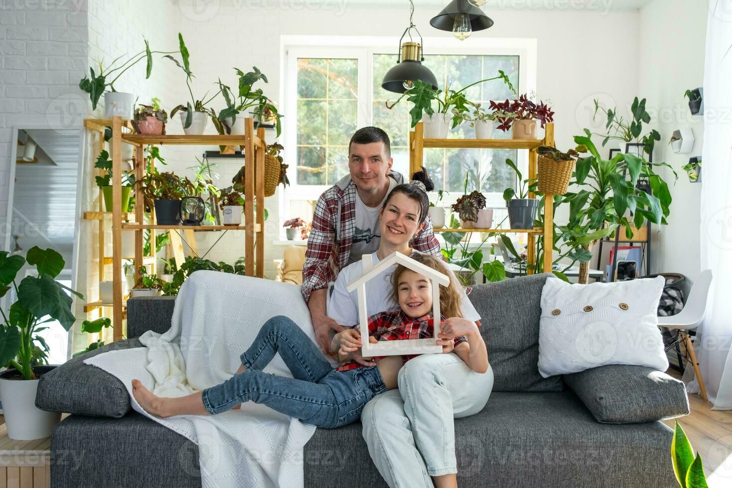 tradicional família com criança estão desfrutando Novo lar, sentado em a sofá e segurando a teto. hipoteca, seguro e proteção, comprando e comovente para dentro seu próprio lar, verde casa com em vaso plantar foto