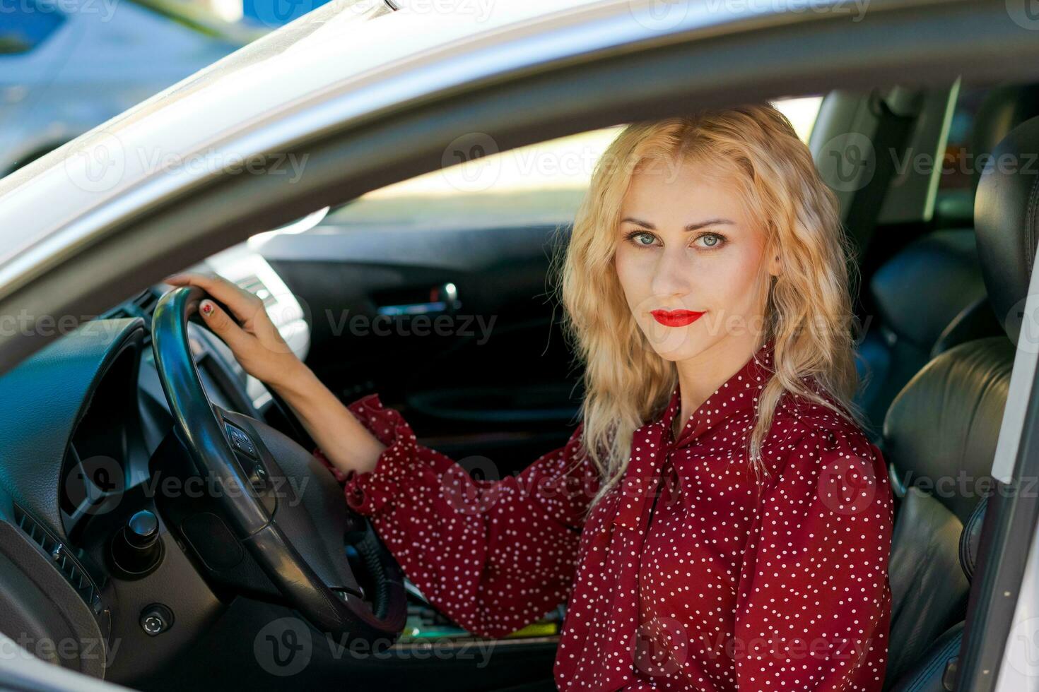 lindo bem sucedido Loiras mulher dentro uma vermelho vestir sentado dentro uma carro atrás a roda foto