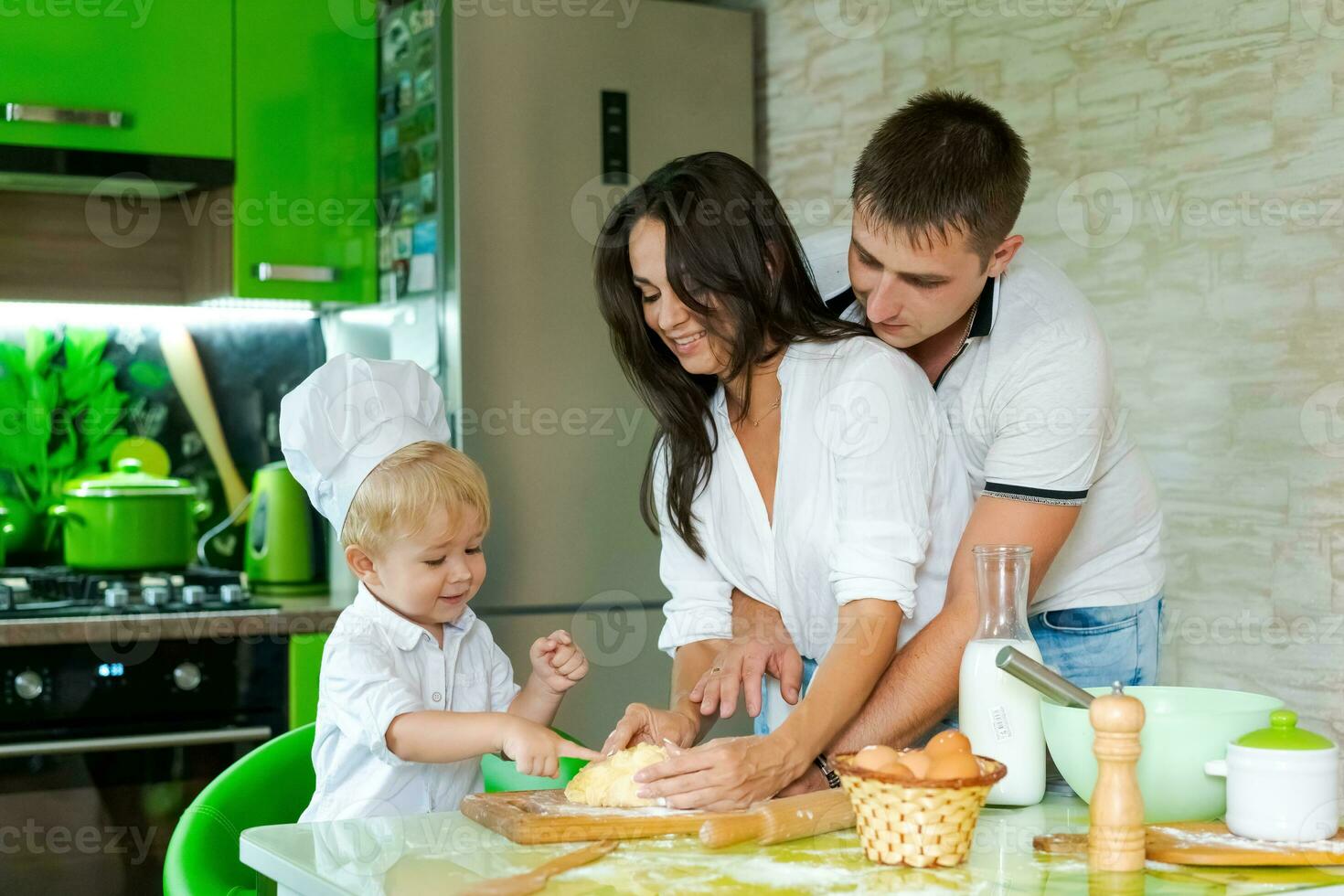 feliz família mãe e pequeno filho e Papai estão preparando massa dentro cozinha às mesa. produtos para massa estão em mesa foto