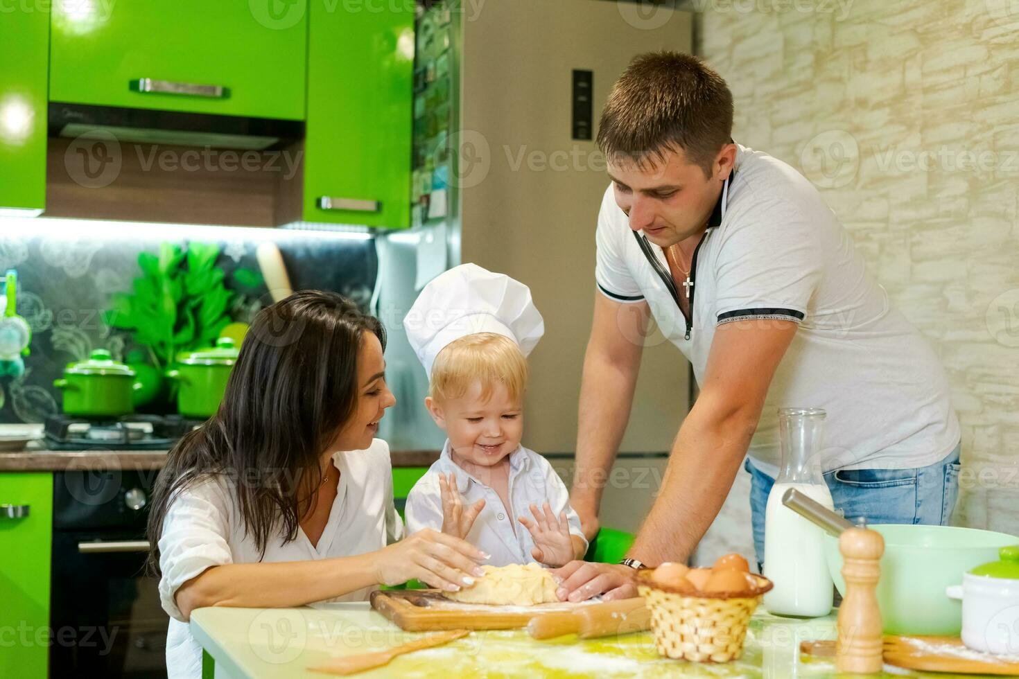 feliz família mãe e pequeno filho e Papai estão preparando massa dentro cozinha às mesa. produtos para massa estão em mesa foto