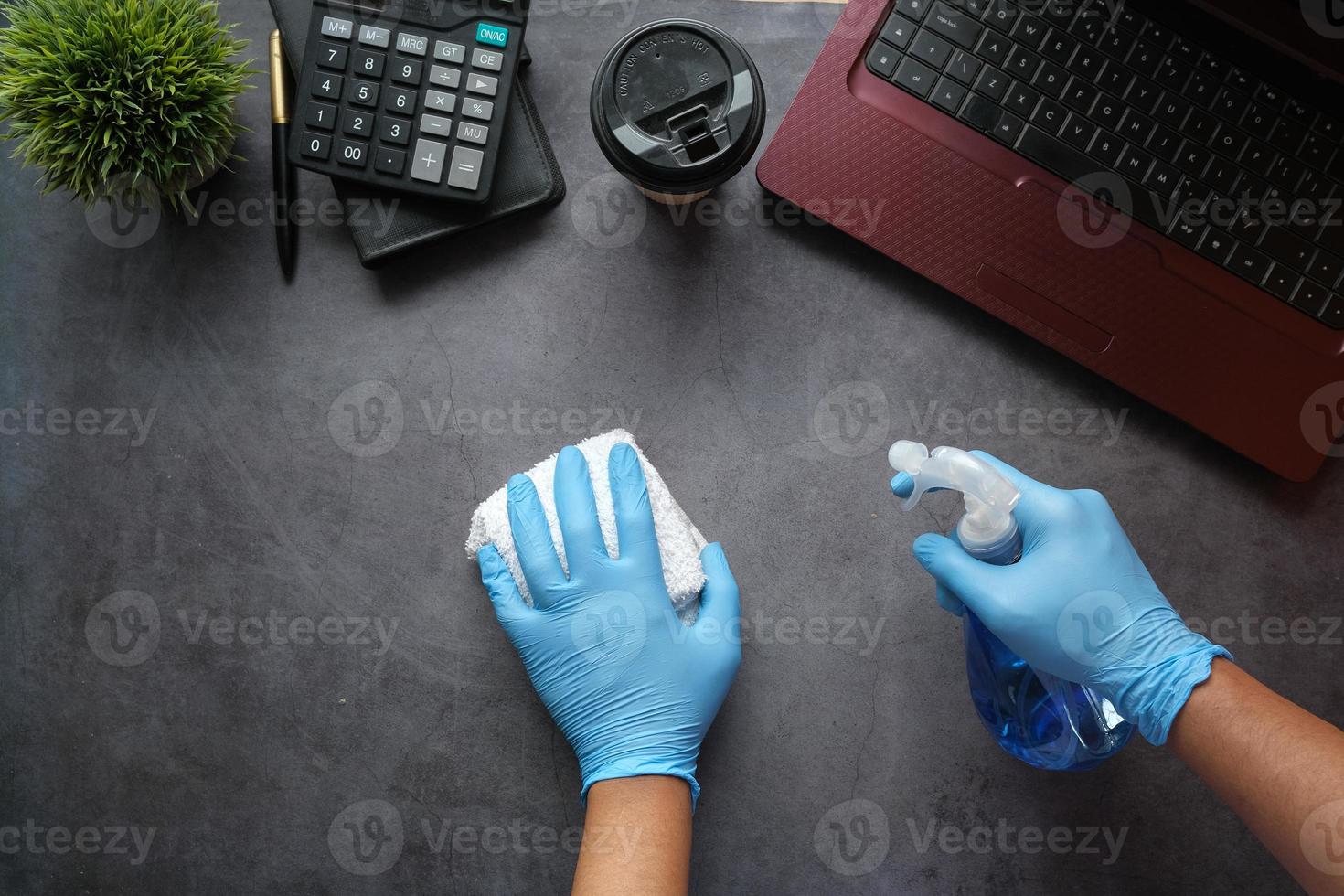 mãos com luvas de borracha azuis segurando um frasco de spray para limpar a mesa do escritório foto