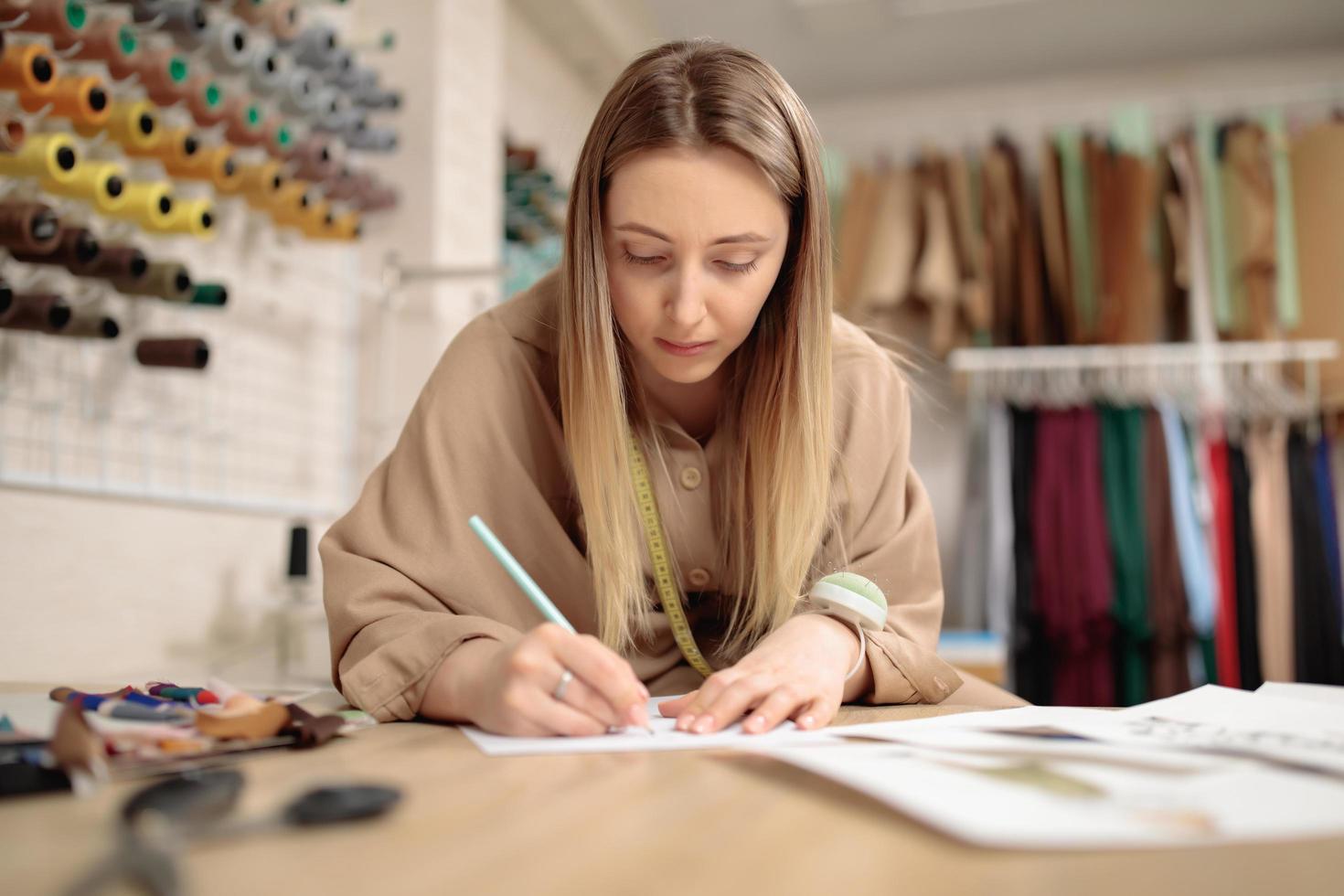 designer de moda jovem artista feminina está desenhando um esboço na mesa no estúdio moderno. pequeno alfaiate foto