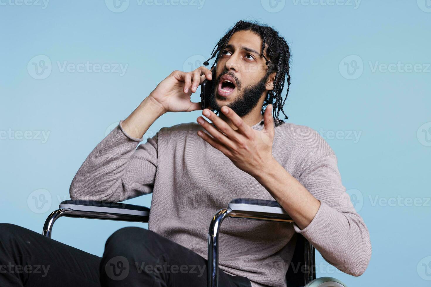 emocional irritado árabe homem com incapacidade falando em Smartphone enquanto sentado dentro cadeira de rodas. Bravo pessoa com fisica prejuízo fazer gestos enquanto conversando em Móvel telefone foto