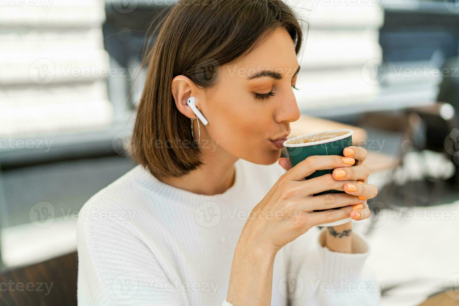 feliz curto cabelos mulher desfrutando cappucino dentro cafeteria. vestindo acolhedor branco suéter. ouvindo favorito música de fones de ouvido. foto