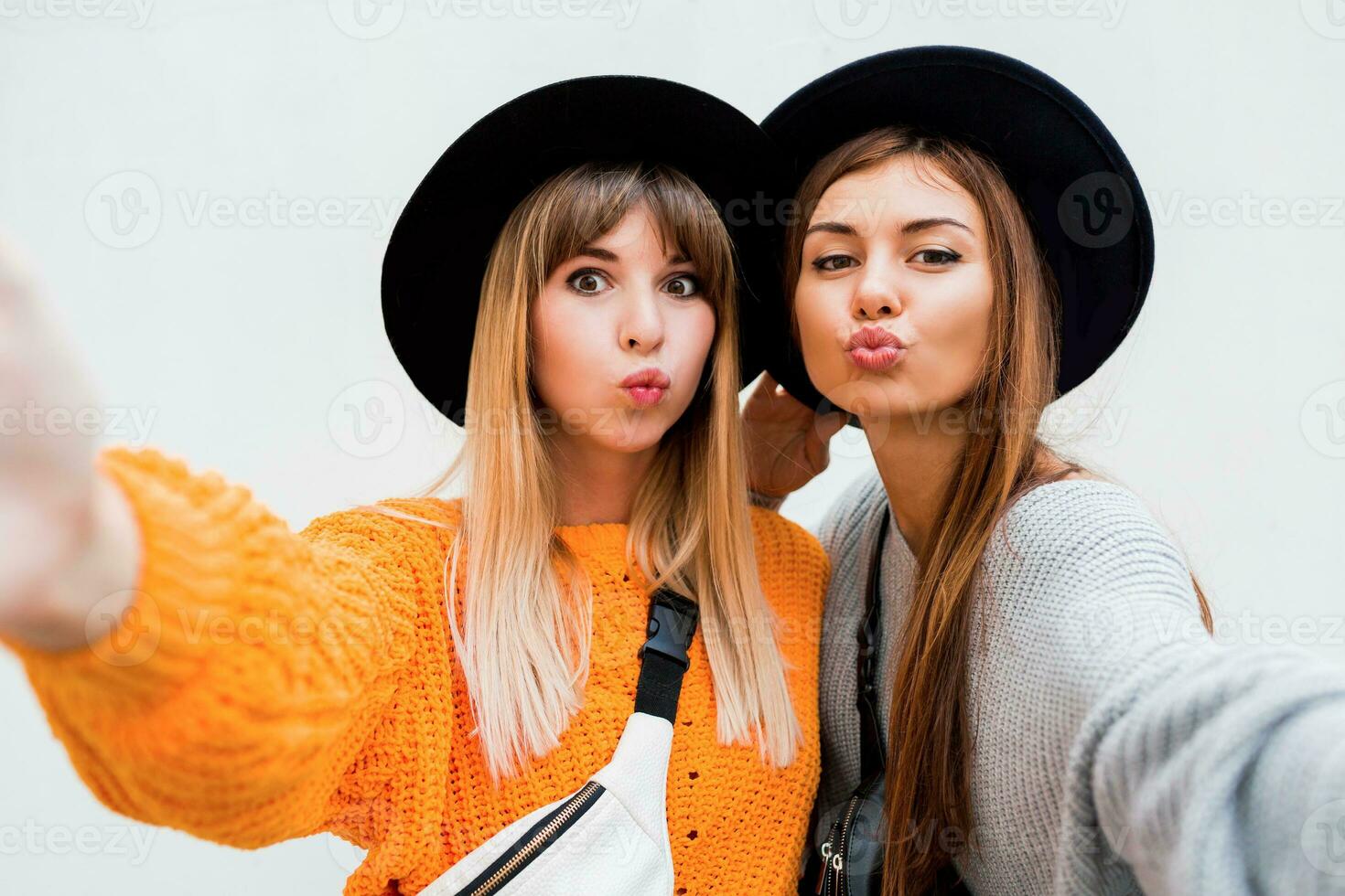 amizade, felicidade e pessoas conceito. dois sorridente meninas sussurrando fofoca em branco fundo. laranja suéter, Preto semelhante chapéus. foto