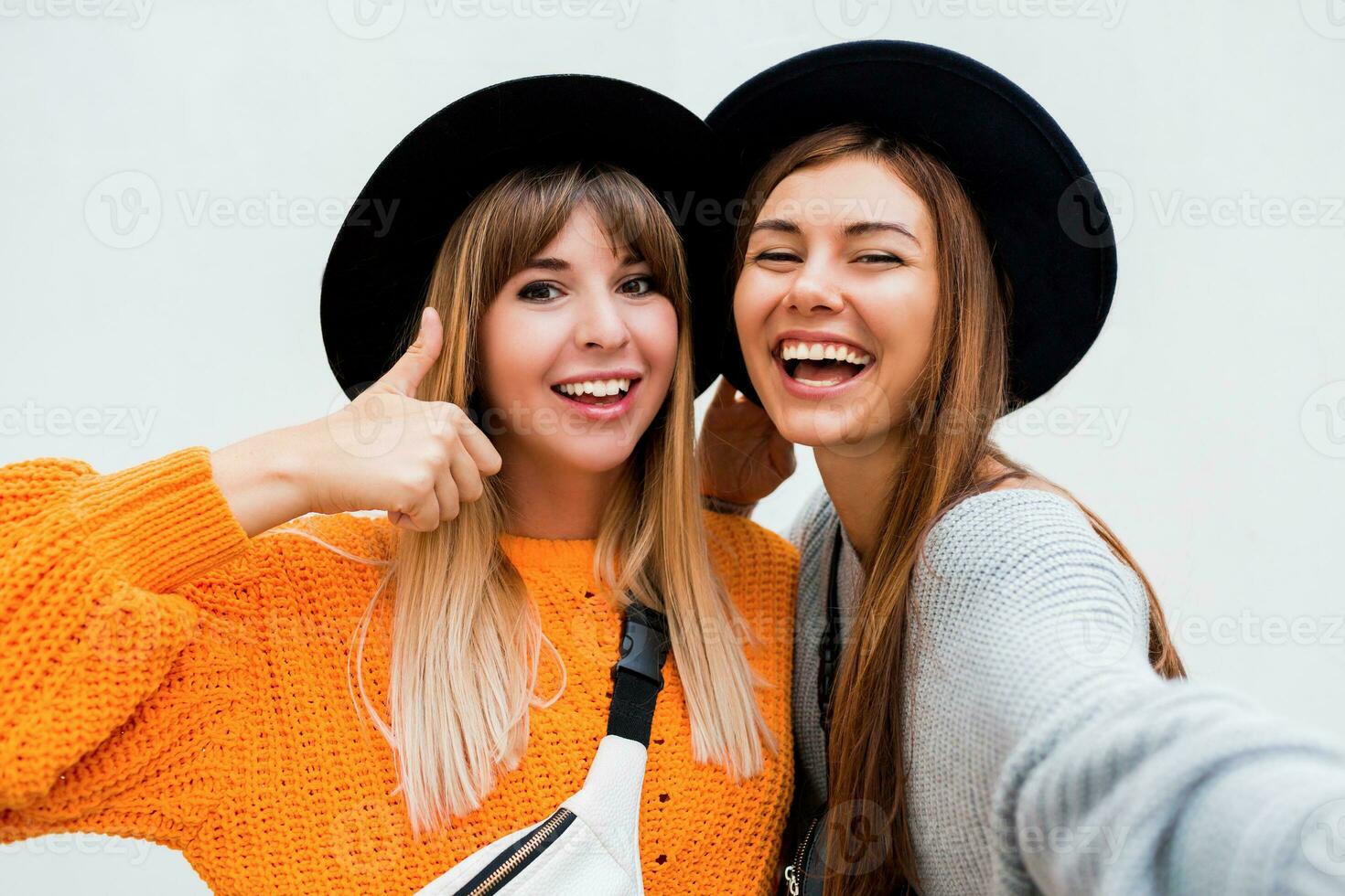 amizade, felicidade e pessoas conceito. dois sorridente meninas sussurrando fofoca em branco fundo. laranja suéter, Preto semelhante chapéus. foto
