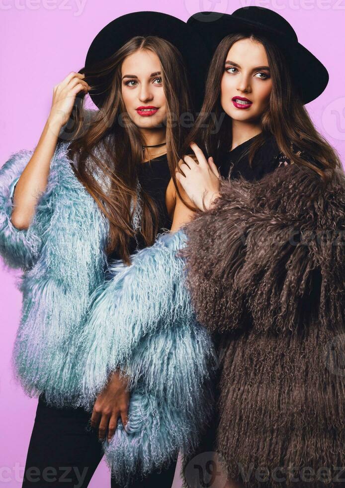 positivo moda retrato do dois garotas, melhor amigos posando interior em brilhante Rosa fundo vestindo inverno à moda fofo casaco, Preto casual chapéu. elegante roupas. irmãs andando. foto