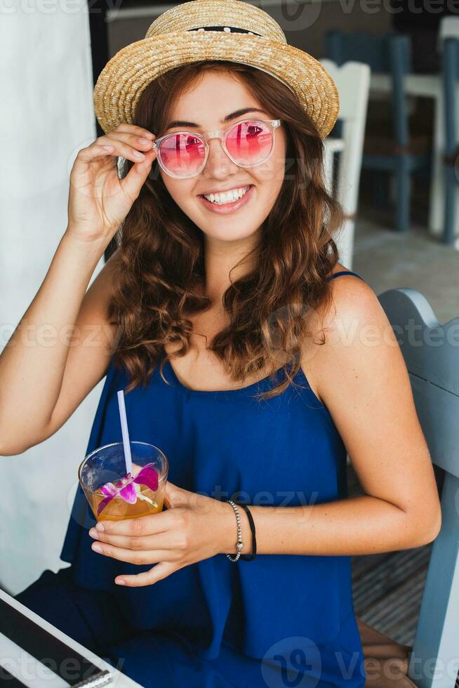 atraente jovem mulher dentro azul vestir e Palha chapéu vestindo Rosa oculos de sol bebendo coquetéis foto