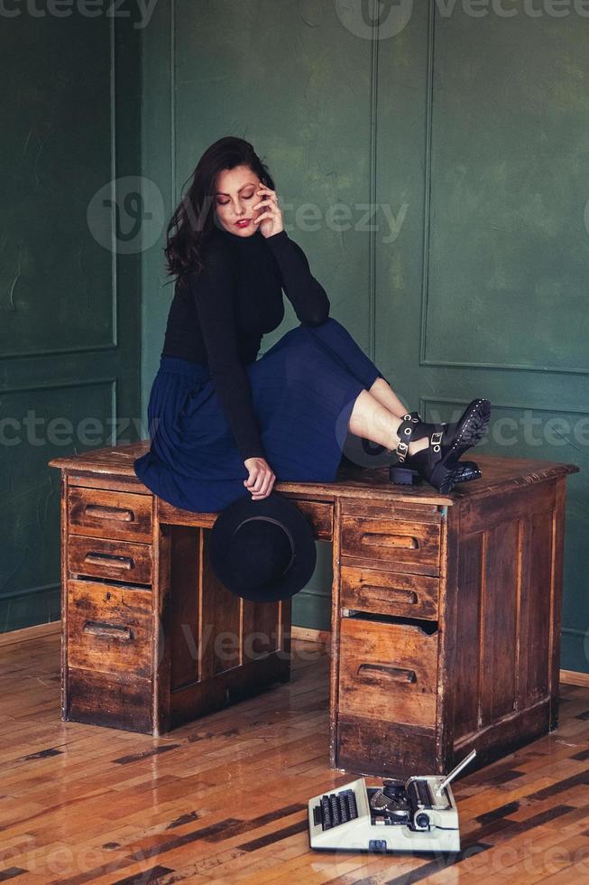 Mulher bonita com um chapéu sentada perto de uma mesa de carvalho com uma máquina de escrever vintage foto