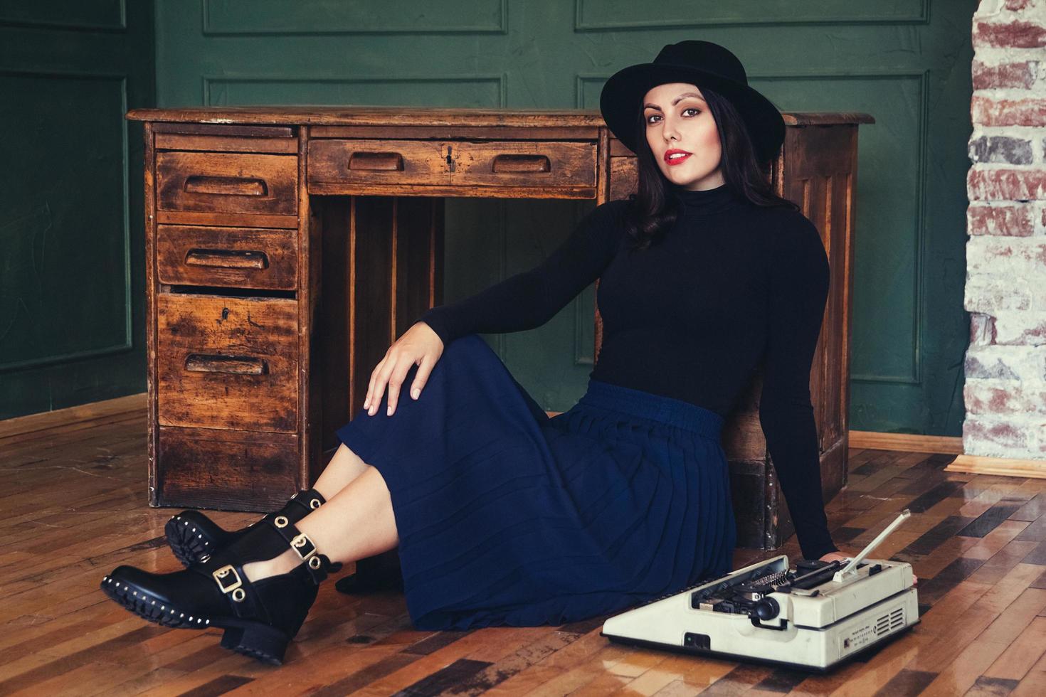 Mulher bonita com um chapéu sentada perto de uma mesa de carvalho com uma máquina de escrever vintage foto