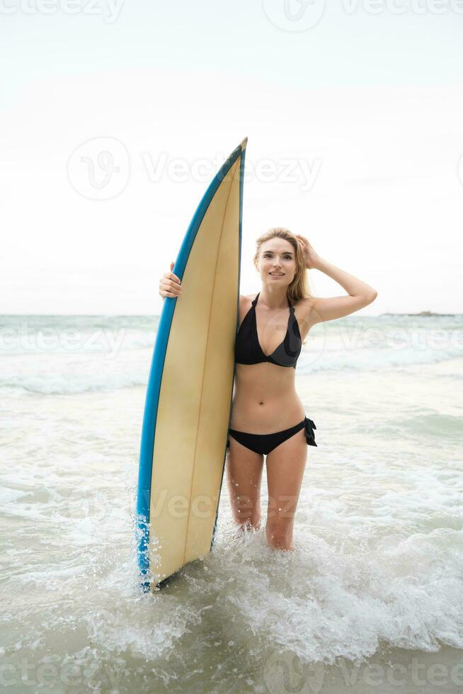 retrato do sorridente jovem mulher em pé com prancha de surfe às a de praia foto