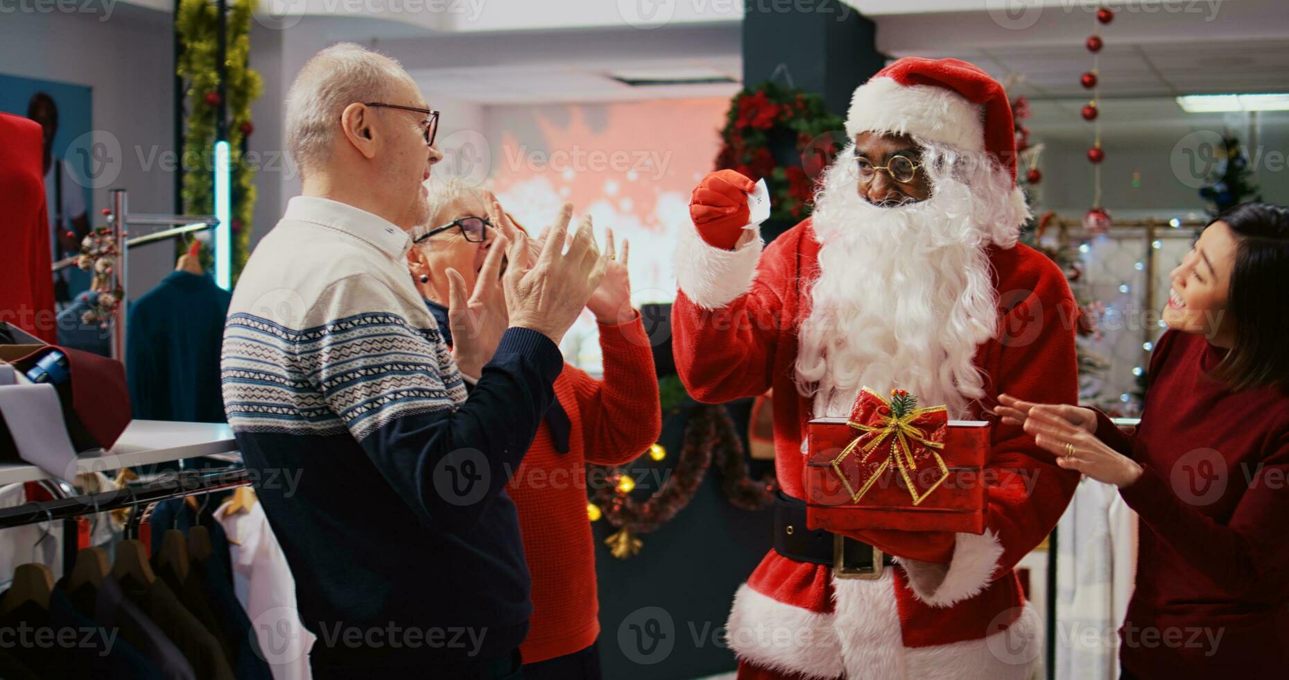 varejo assistente vestindo santa claus terno dentro feriado adornar moda butique, segurando presente caixa agindo Como sorteio bilhetes recipiente, desenhando aleatória Senior mulher entre feliz clientes foto