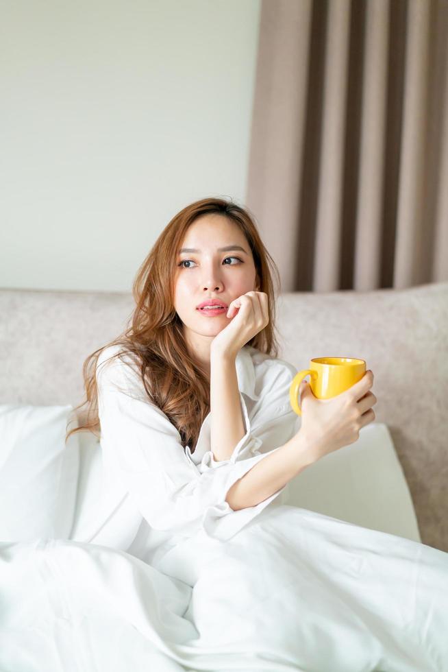 retrato linda mulher asiática acordar e segurando uma xícara de café ou caneca na cama pela manhã foto