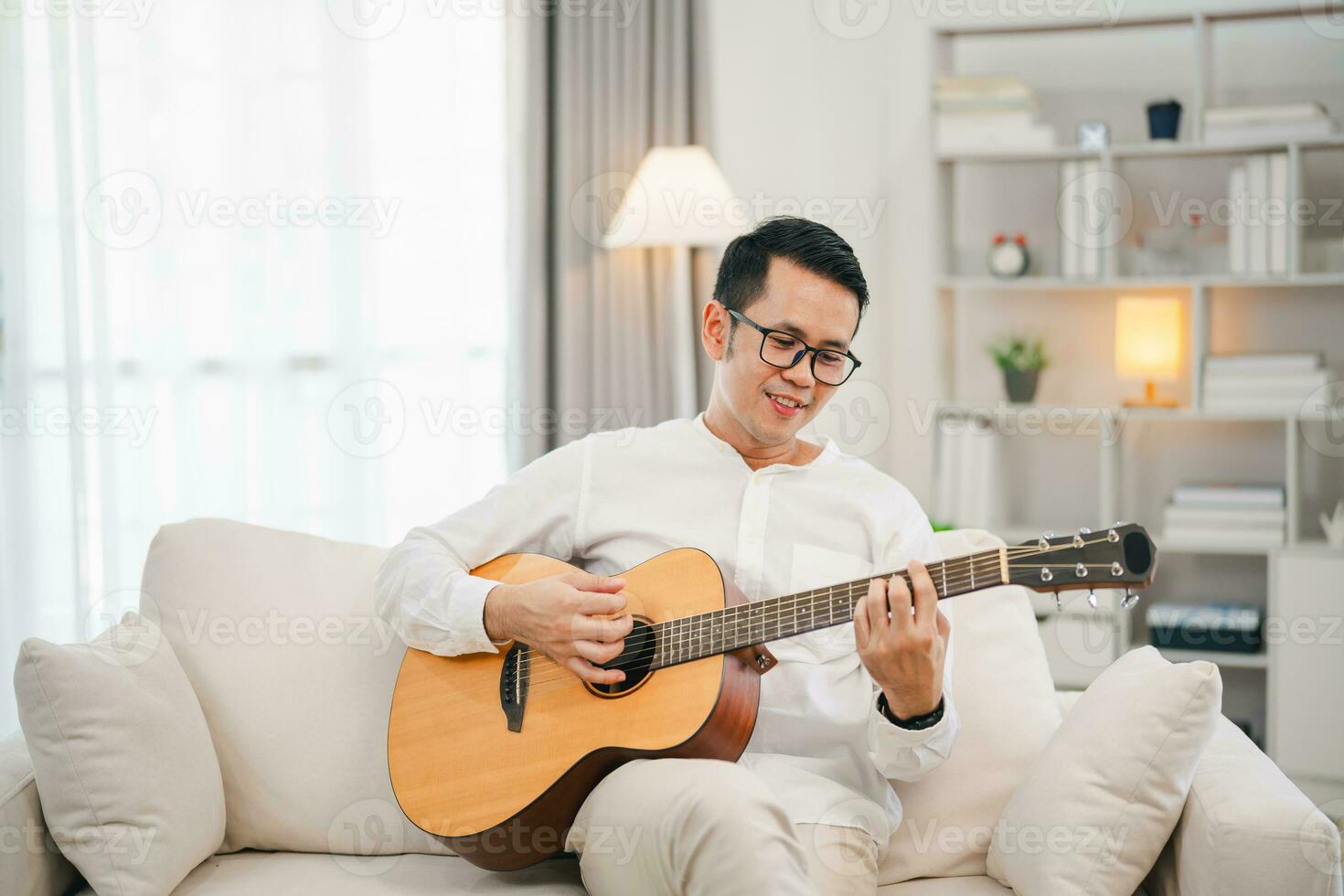 ásia homem vestindo óculos e jogando guitarra enquanto sentado em sofá dentro a vivo quarto às lar. ásia homem escrevendo música enquanto jogando guitarra às lar. compor música música conceito. foto