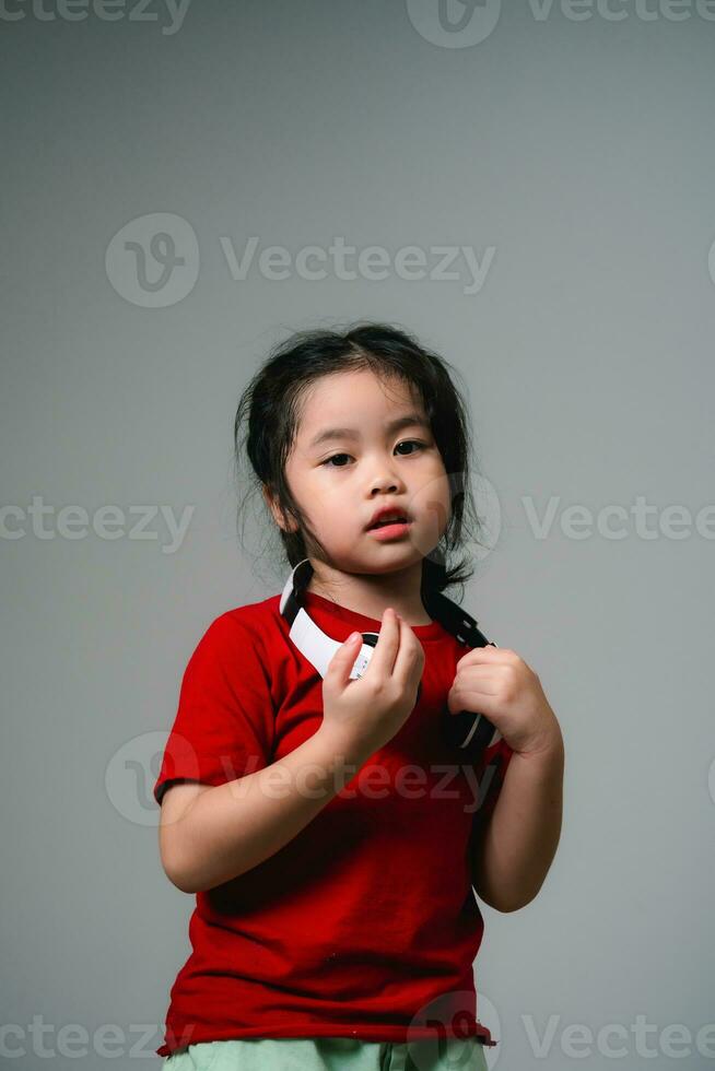 alegre feliz ásia crianças menina com Preto cabelo ouvindo para música canções dentro fones de ouvido em uma cinzento fundo. retrato fofa menina advertência vermelho camisa e olhando às Câmera estúdio iluminação. foto