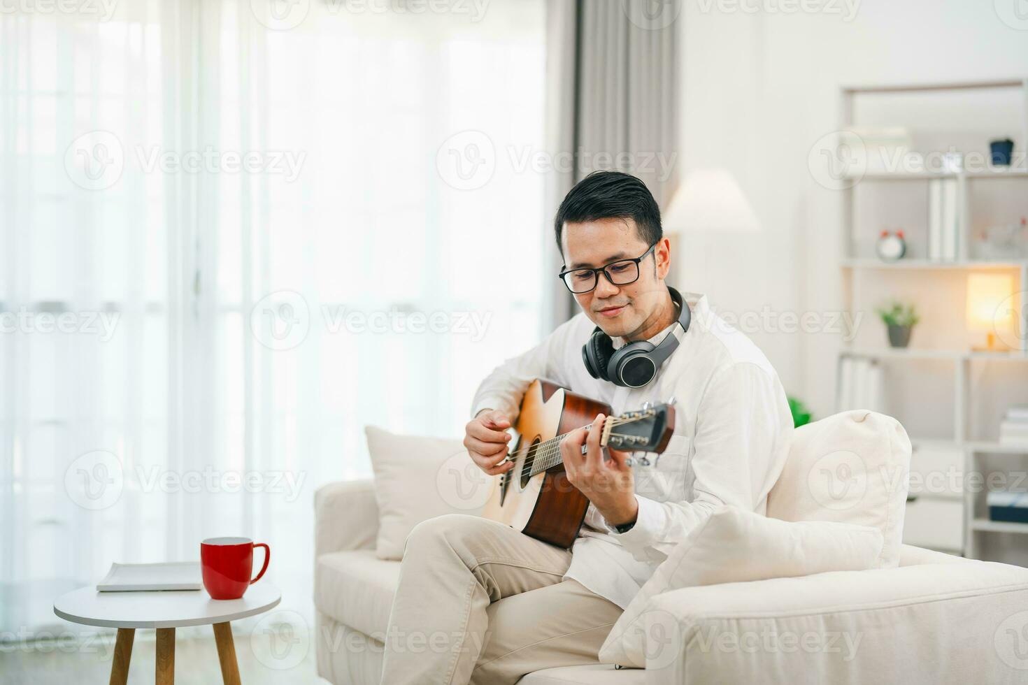 ásia homem vestindo óculos e fone de ouvido jogando guitarra enquanto sentado em sofá dentro a vivo quarto às lar. ásia homem escrevendo música enquanto jogando guitarra às lar. compor música música conceito. foto