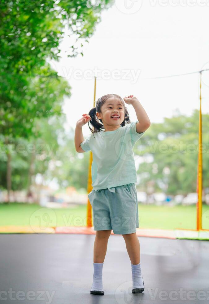 ásia criança menina é pulando em trampolim em Parque infantil fundo. feliz rindo criança ao ar livre dentro a Jardim em verão período de férias. saltar Alto em trampolim. atividade crianças dentro a Jardim da infância escola. foto
