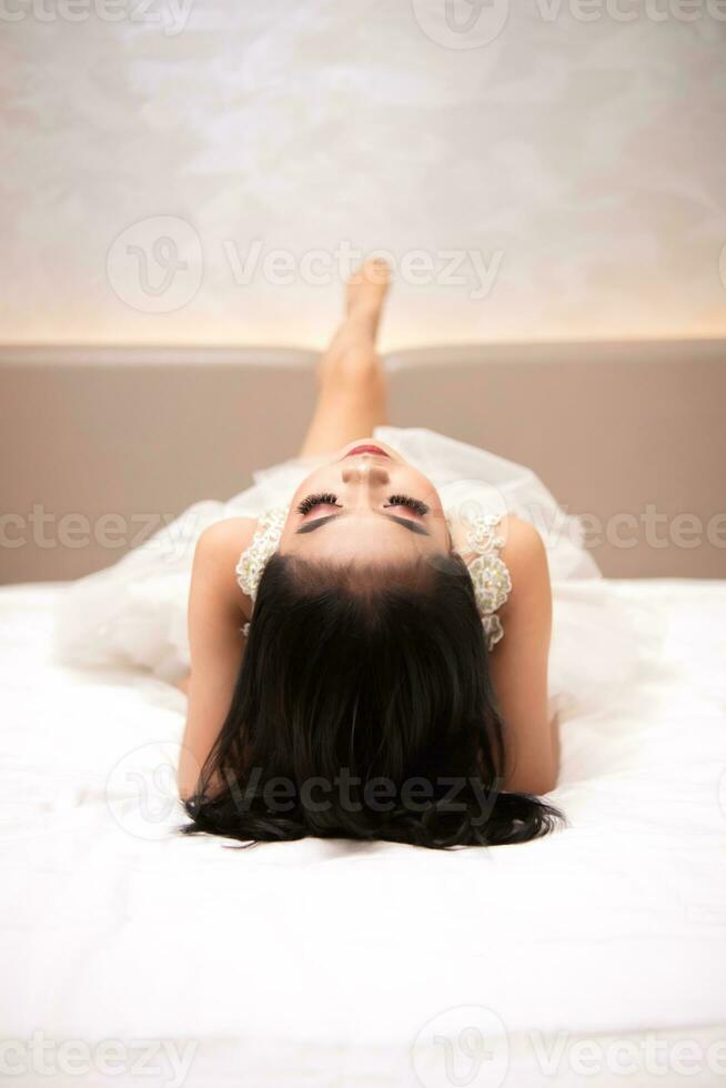 a ásia mulher dentro uma branco vestir é deitado em uma branco cama com dela Preto cabelo solto dentro uma hotel foto