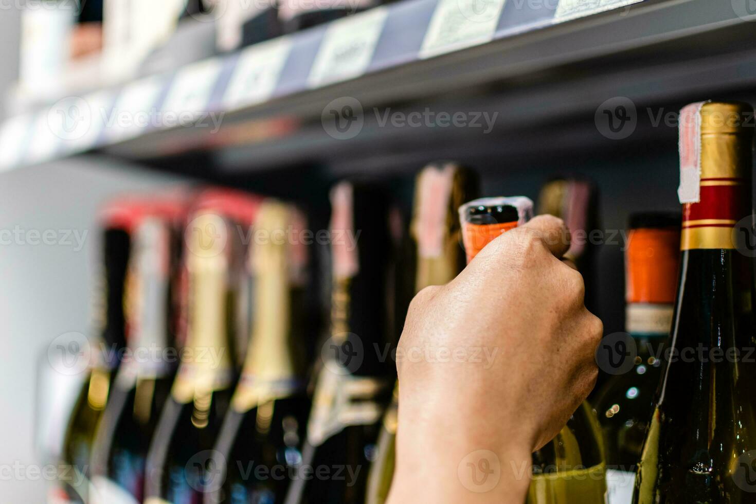 fêmea cliente contenção, escolhendo uma vermelho vinho garrafa rótulo dentro uma departamento loja. mulher detém uma garrafa do vinho às supermercado álcool seção. estante é cheio do alcoólico bebidas. borrado fundo foto