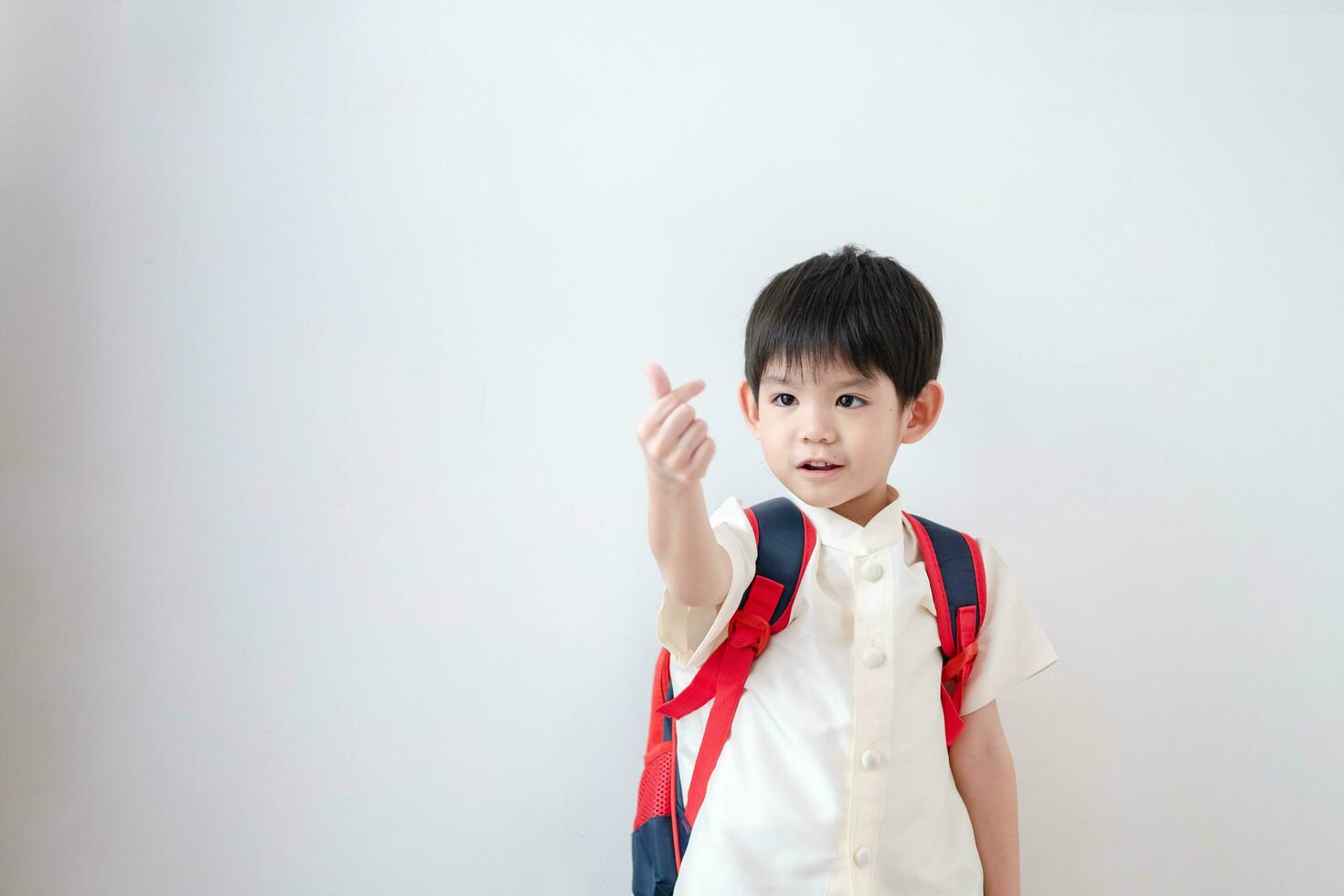 ásia Garoto vestindo tradicional tailandês roupas, em pé com uma escola saco dentro mão, fazer uma mão mini coração, preparando para ir para escola. em uma branco fundo foto