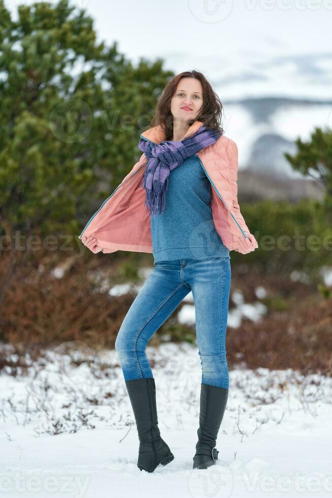 jovem mulher carrinhos dentro inverno floresta perto conífero árvore, olhando às Câmera. morena mulher com grandes cabelo, vestido dentro bege jaqueta, azul jeans, de pescoço alto chuteiras foto