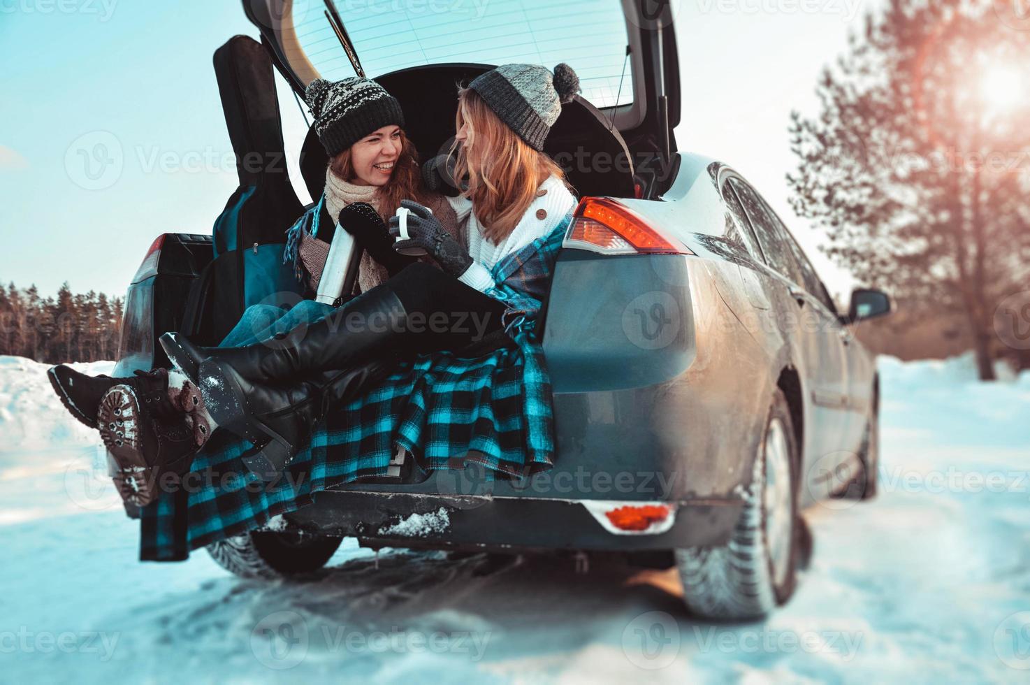 amigos felizes na floresta de inverno. duas meninas sentadas no porta-malas bebendo café foto