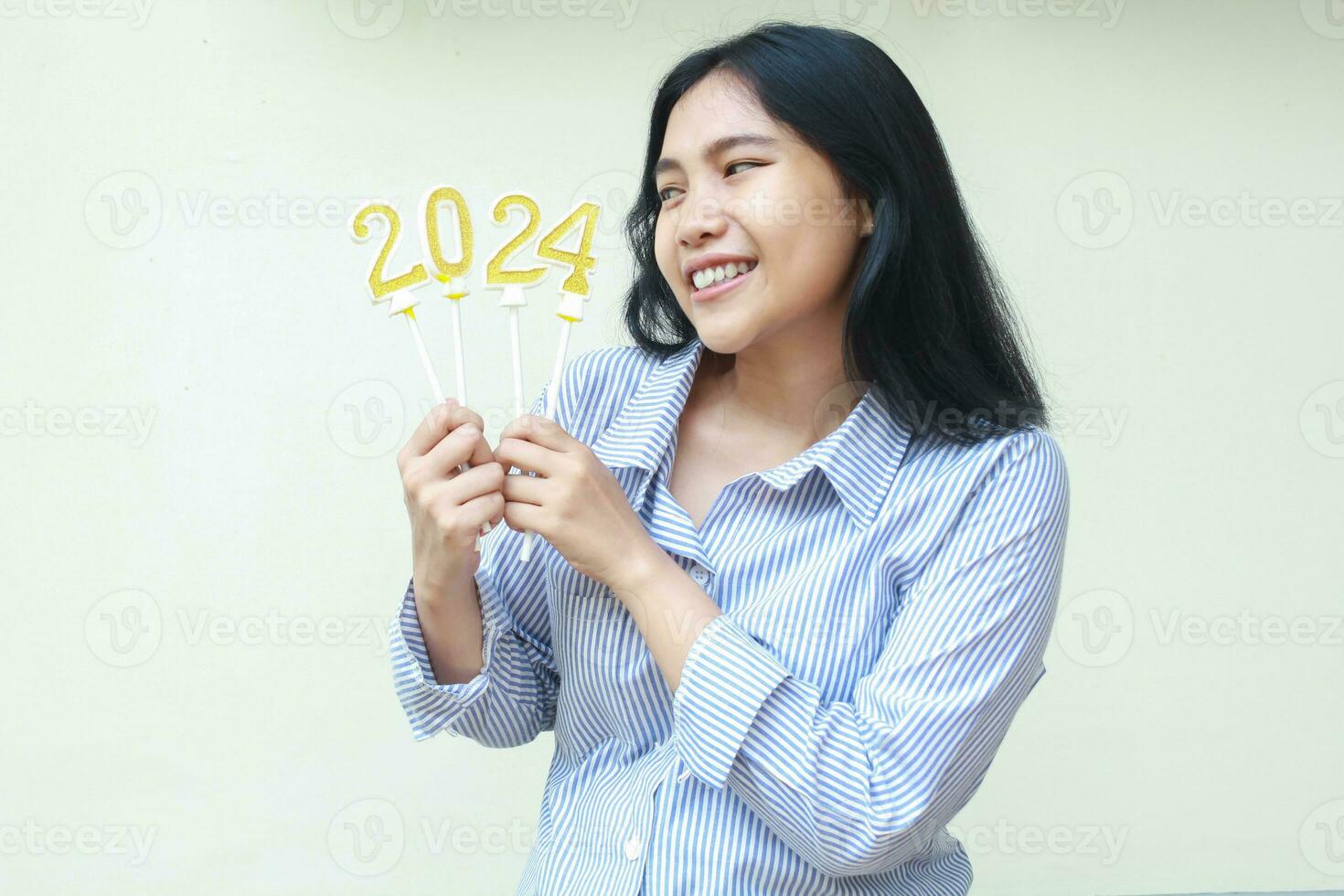 animado ásia jovem o negócio mulher a comemorar Novo anos véspera olhando para dourado velas números 2024 aguarde em mão vestindo listras azul camisa isolado em branco fundo foto