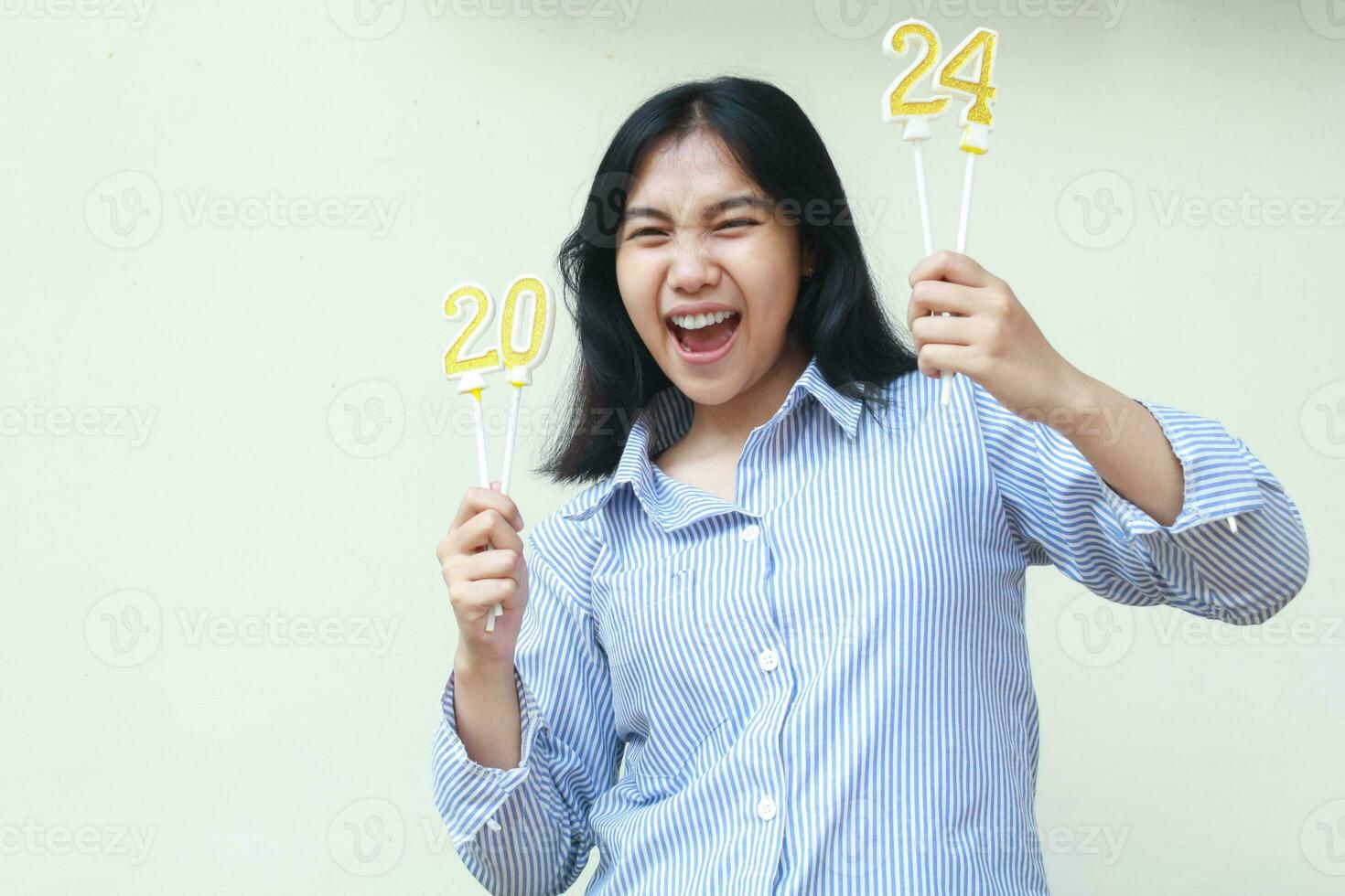 animado ásia jovem mulher rir e grito a comemorar Novo anos véspera de segurando dourado velas vestindo azul listras camisa olhando às Câmera isolado em branco fundo foto