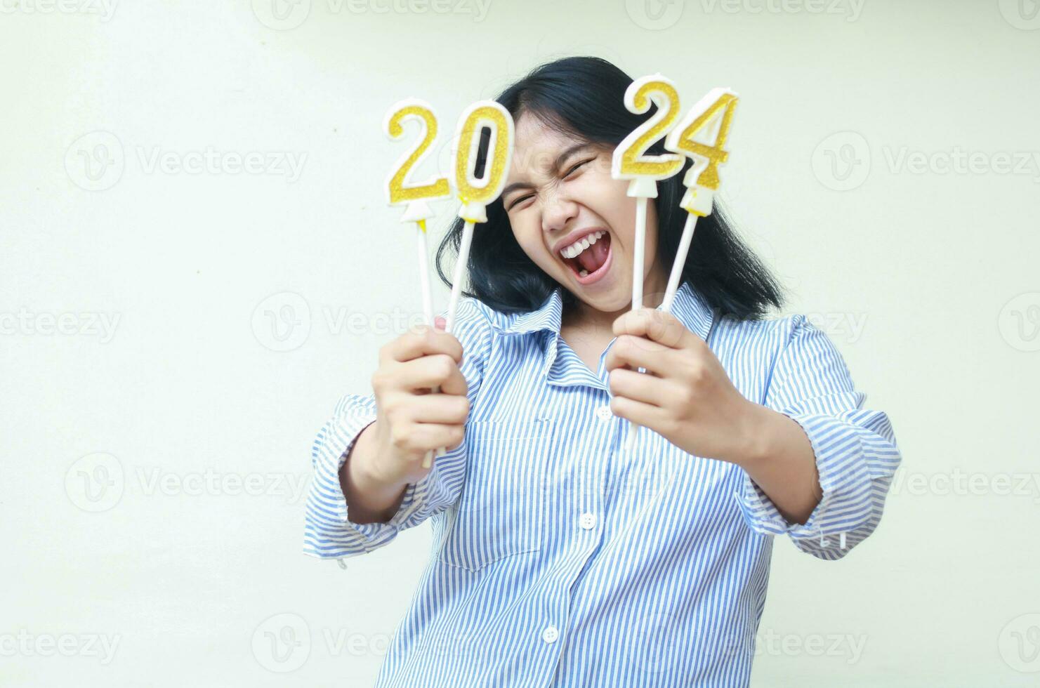 animado jovem ásia mulher gritando para Câmera com levantando braço segurando 2024 vela número, isolado, fêmea colega de trabalho vestindo azul listras camisa casual, Novo anos véspera conceito foto