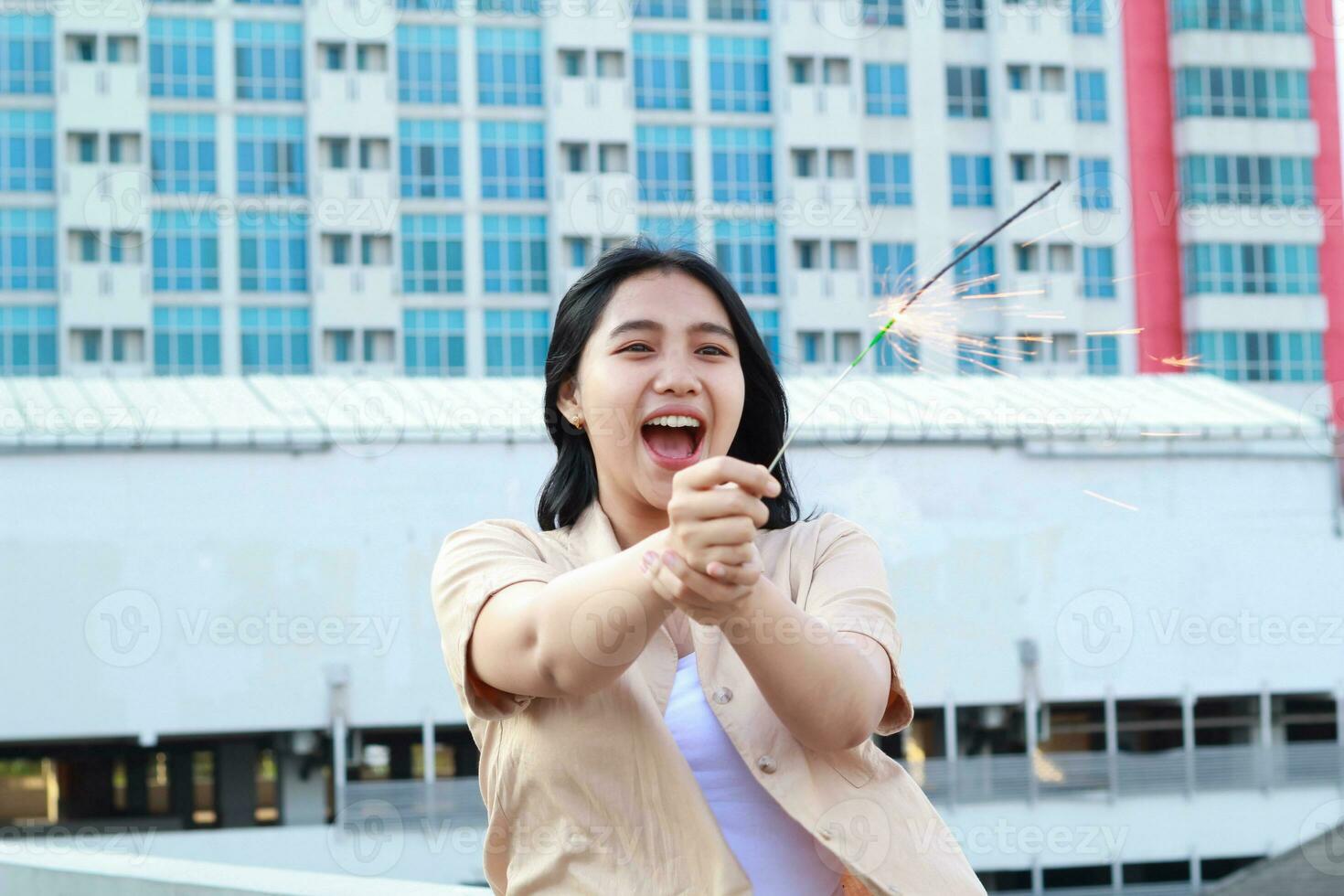 lindo ásia jovem mulher segurando estrelinhas fogo de artifício e rindo dentro Novo ano véspera celebração em pé dentro cobertura topo ao ar livre com urbano construção fundo foto