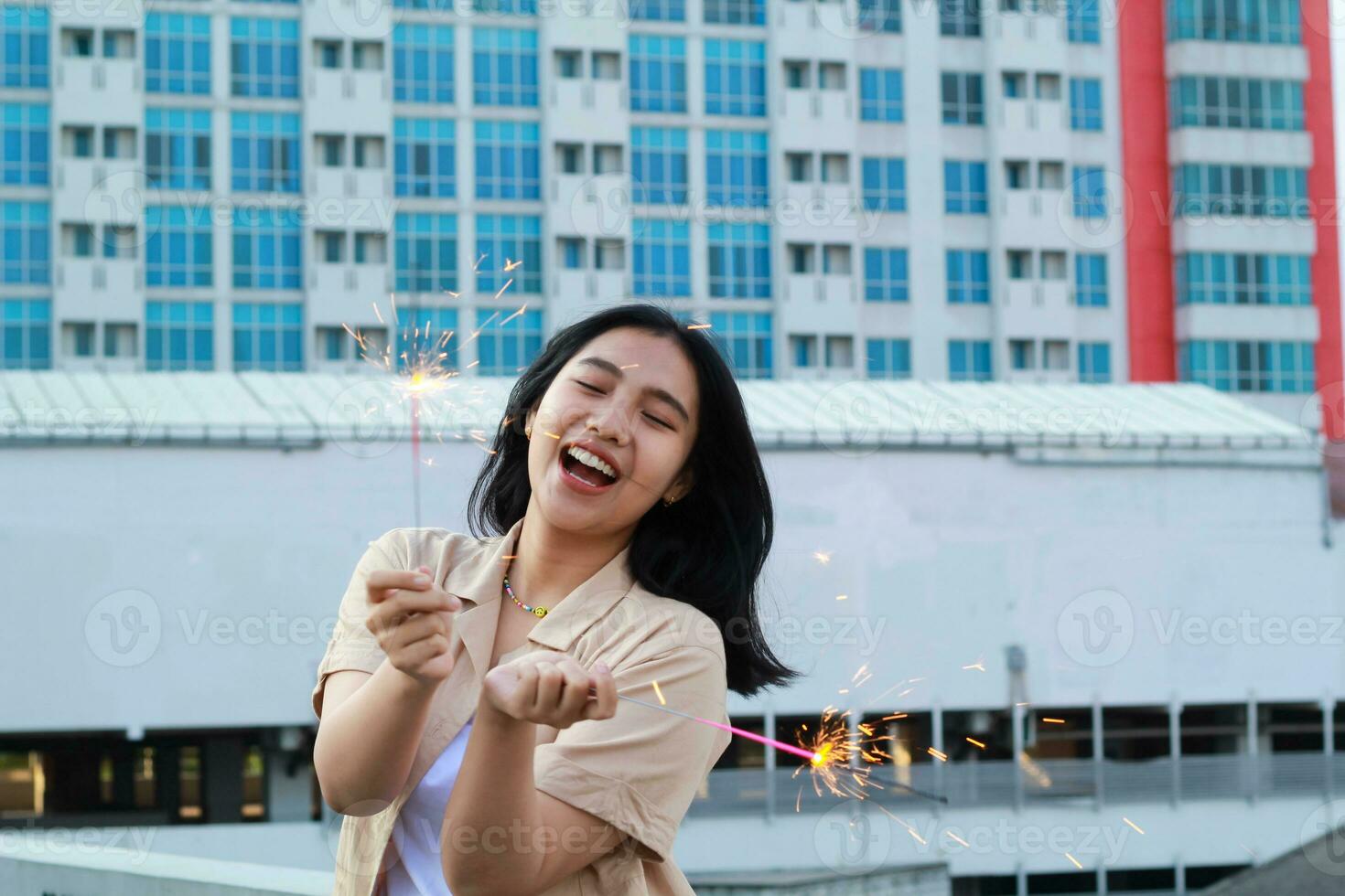 despreocupado ásia jovem mulher segurando diamante comemoro Novo ano véspera com rir e dança dentro telhado apartamento ao ar livre com cidade construção fundo foto