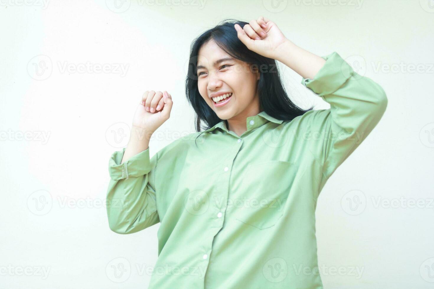 brincalhão sucesso Sombrio cabelo ásia mulher rindo e dançando para comemoro campeão ganhar vitória de levantar punho vestindo verde grande demais camisa em pé sobre isolado branco fundo, olhando às Câmera foto